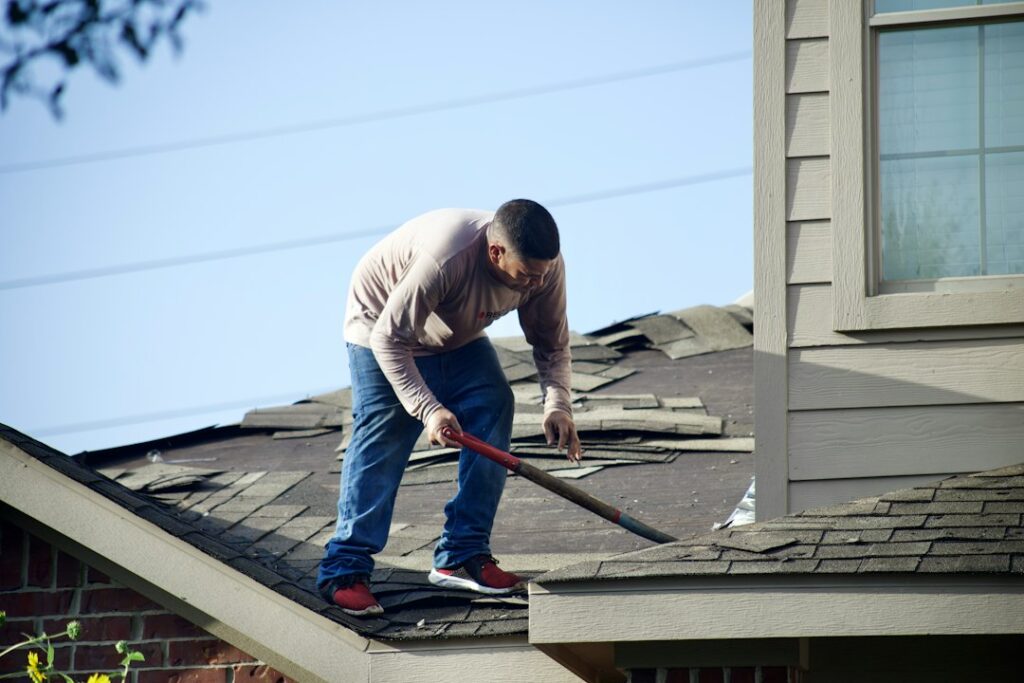 Person examining roof shingles and flashing from a ladder beside a rain gutter on a residential home.