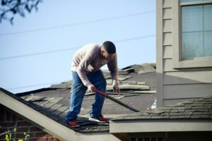 Person examining roof shingles and flashing from a ladder beside a rain gutter on a residential home.